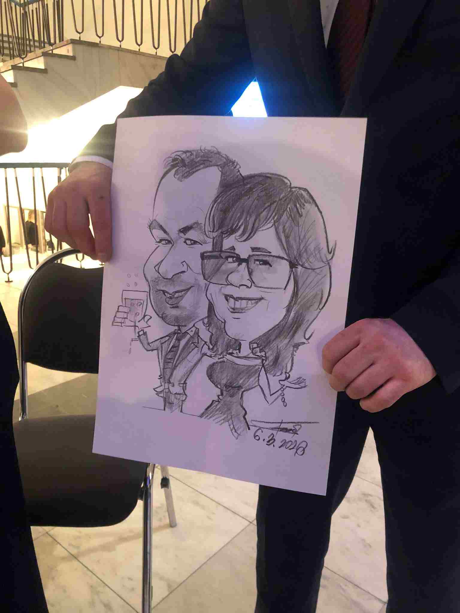 Vtipná karikatúra páru z podujatia, kde muž drží v ruke pohár vína a žena má prehnane veľké oči a široký úsmev.