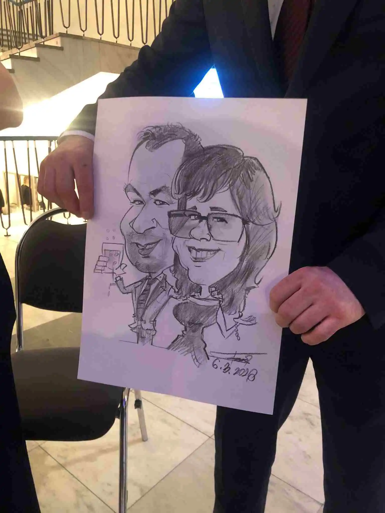 Vtipná karikatúra páru z podujatia, kde muž drží v ruke pohár vína a žena má prehnane veľké oči a široký úsmev.