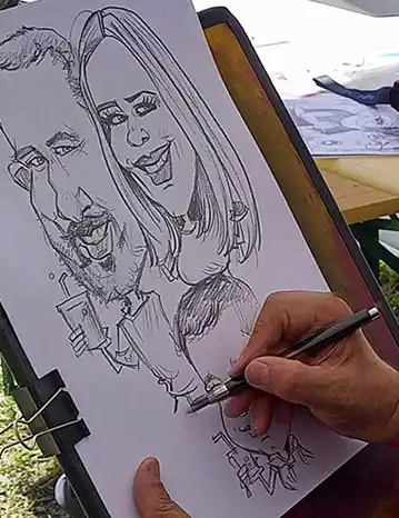 Detailný záber na ruku umelca, ktorý fixkou vytvára vtipný portrét usmievavého páru na formát A4.