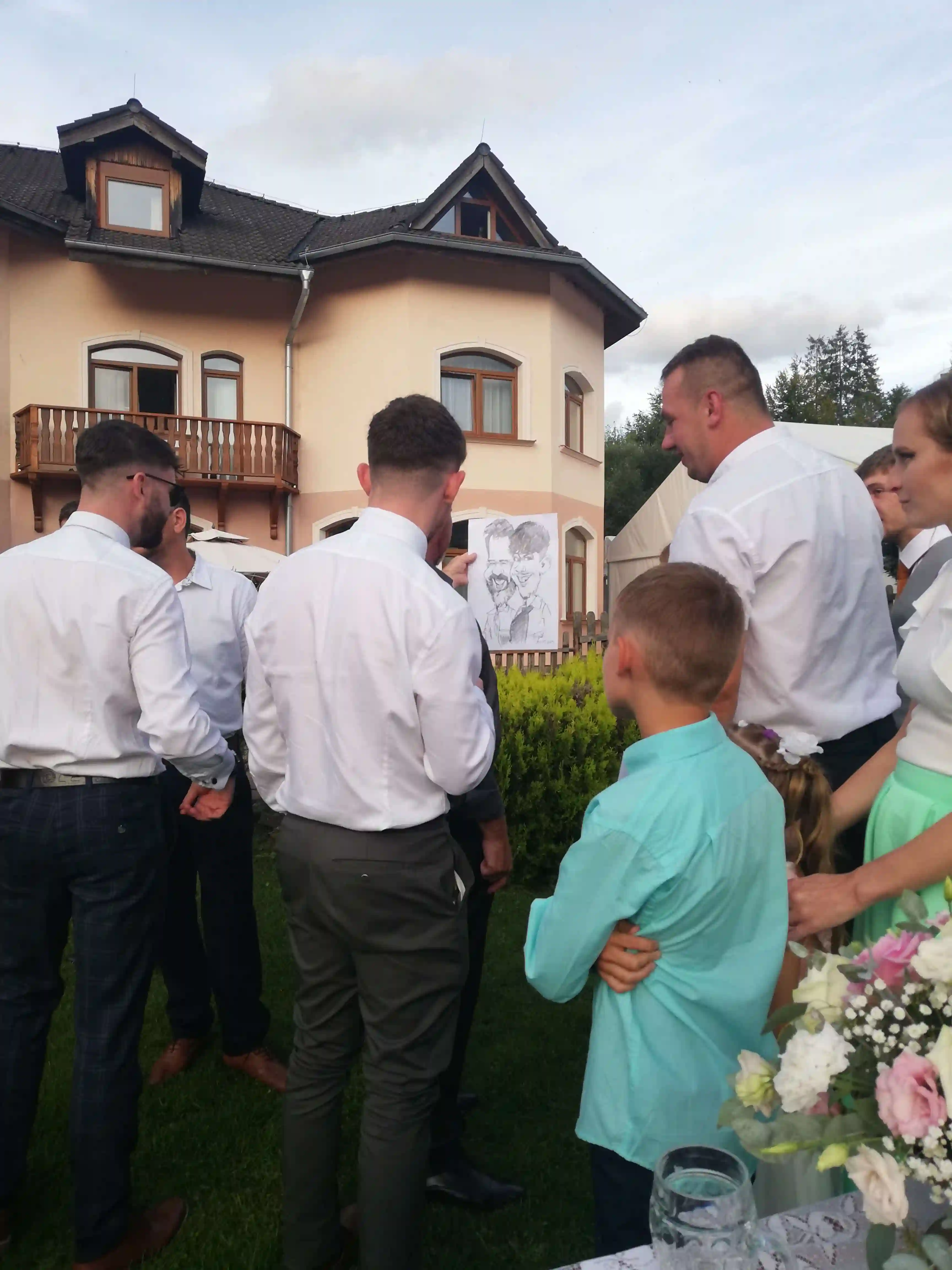 Skupina usmiatych svadobčanov so záujmom sleduje karikaturistu, ktorý práve dokončuje vtipný portrét na papier